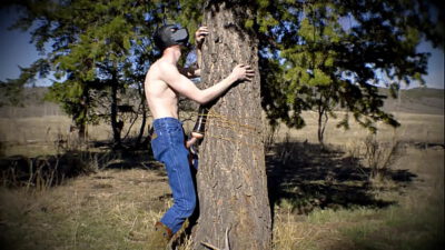 Horny twink in a dark mask fucks a sex toy that is strapped to a tree in a public wilderness.